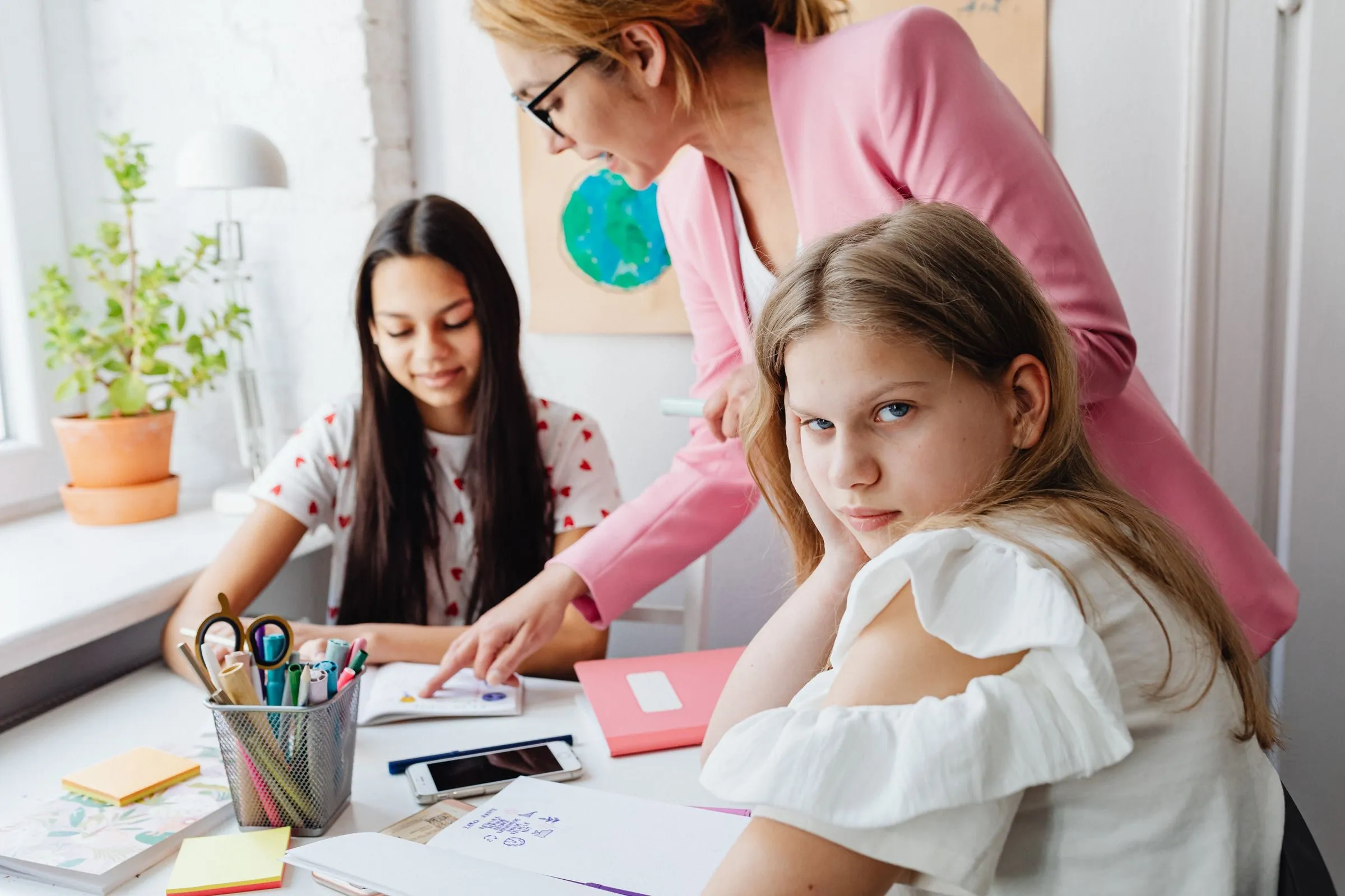 Female teacher helping teenage girls with schoolwork at a desk in a bright classroom setting.